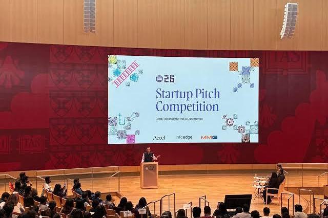 A founder at a podium pitching to a seated audience beneath a large screen